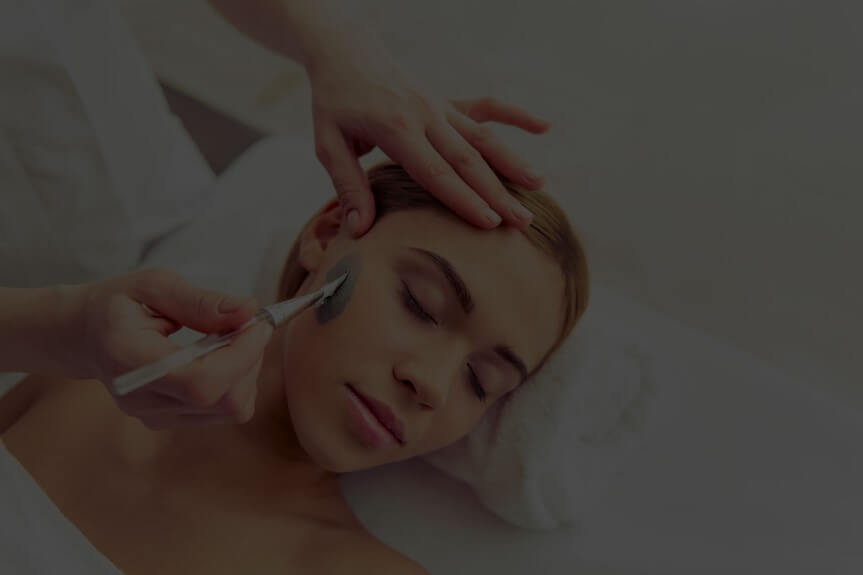 Person lying down with eyes closed, receiving a facial treatment with a brush applying cream.